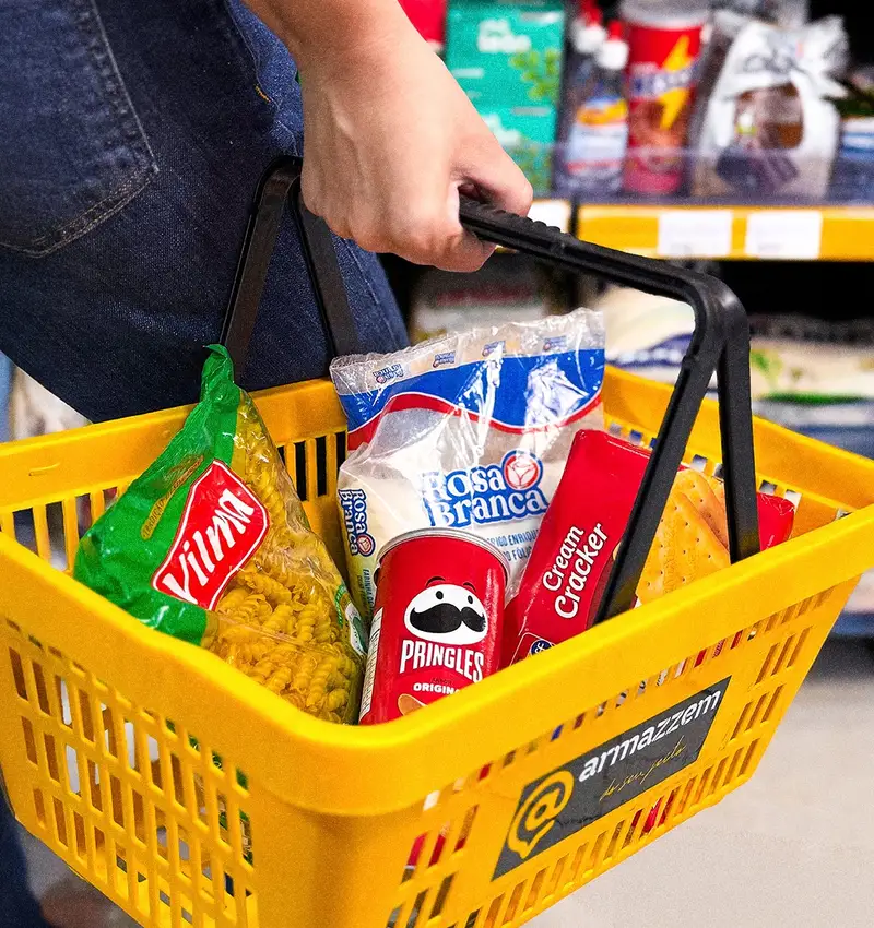 Mão segurando a cesta amarela do Armazzem cheia de produtos, com a loja ao fundo